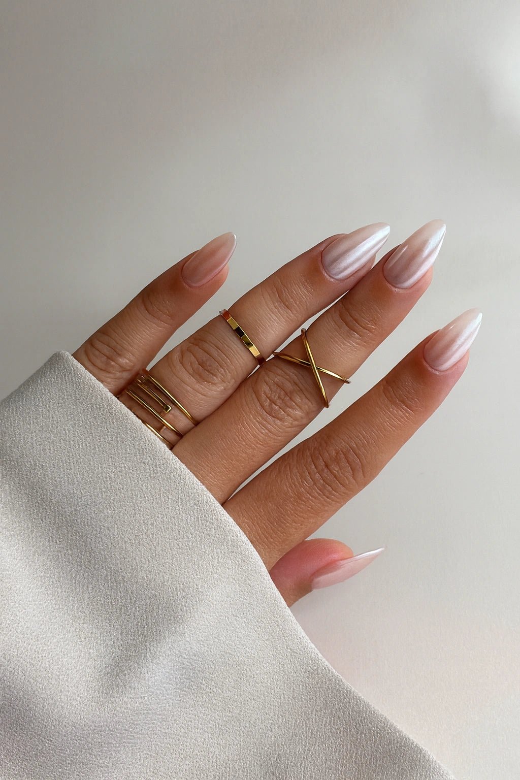 Hand wearing gold rings with light pink chrome fake nails on a neutral background
